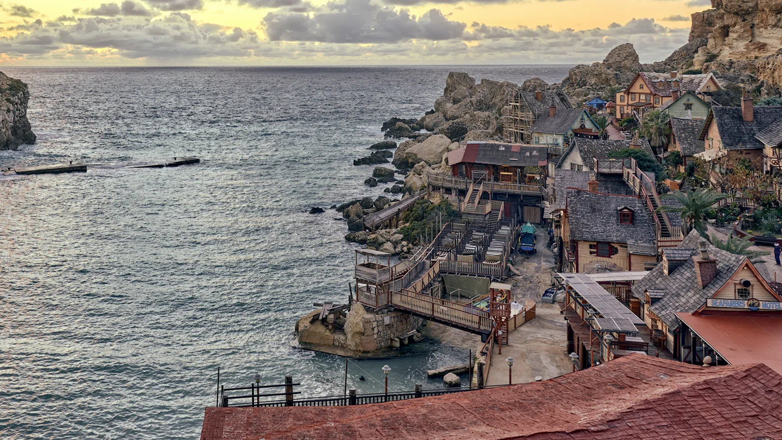 Aerial view of Popeye Village in Anchor Bay, Mellieħa, with weathered wooden buildings clustered along the rocky coastline at golden hour.
